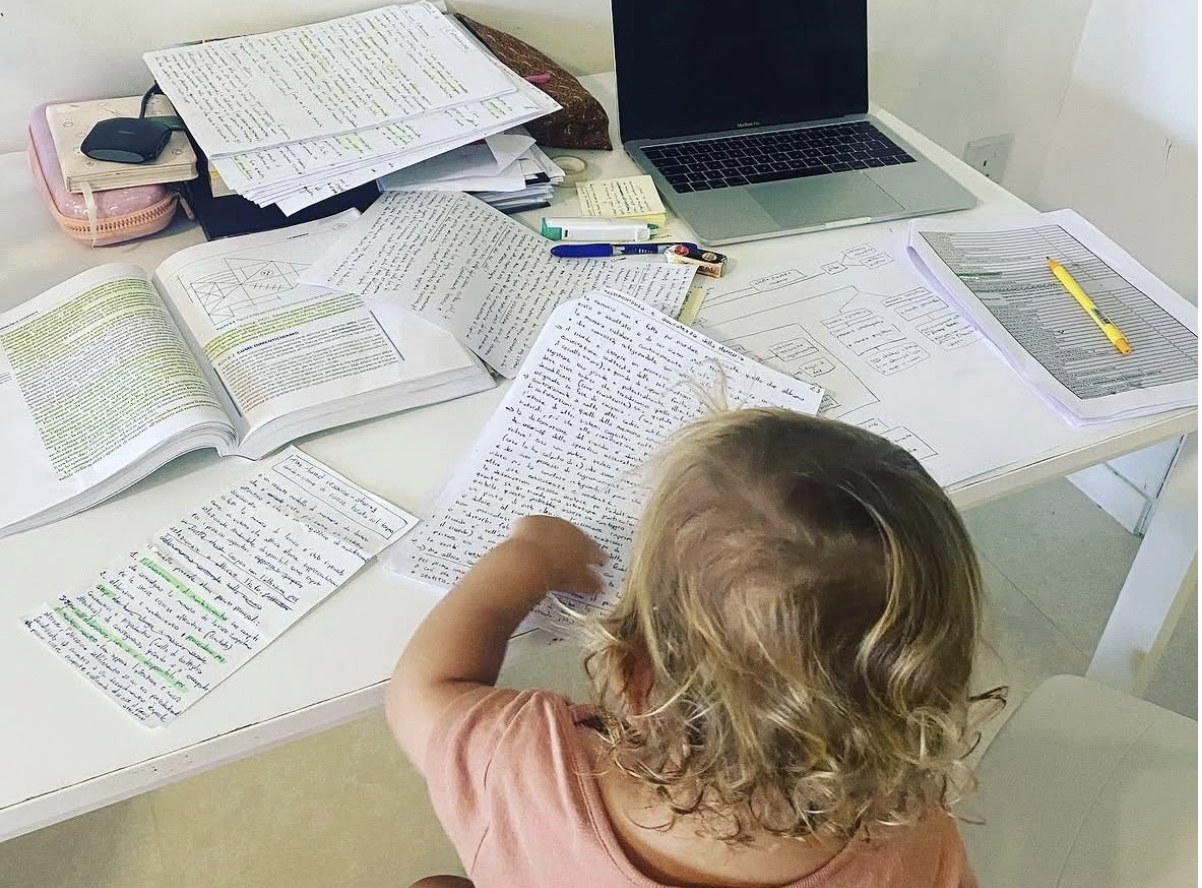 Una scrivania piena di libri e appunti di studio, con una bambina piccola che curiosa tra i fogli